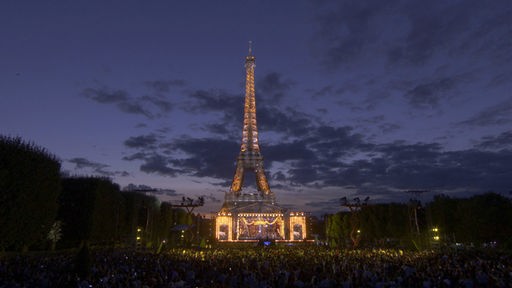 Das Orchestre National de France auf einer Bühne , im Hintergrund der angestrahlte Eifelturm bei Nacht.