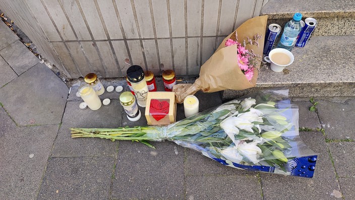 Blumen und Trauerkerzen wurden neben einer Treppe abgelegt. Daneben sind Flaschen und eine Tasse zu sehen.