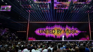 Bunt beleuchtete Bühne mit Schriftzug "United by Music" des Eurovision Song Contest in der Malmö Arena und 2 Personen auf der Bühne, Innenaufnahme mit Publikum