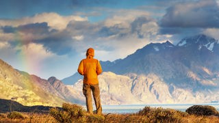Ein von hinten fotografierter Mensch in orangener Outdoorjacke, der auf ein Bergpanorama blickt.